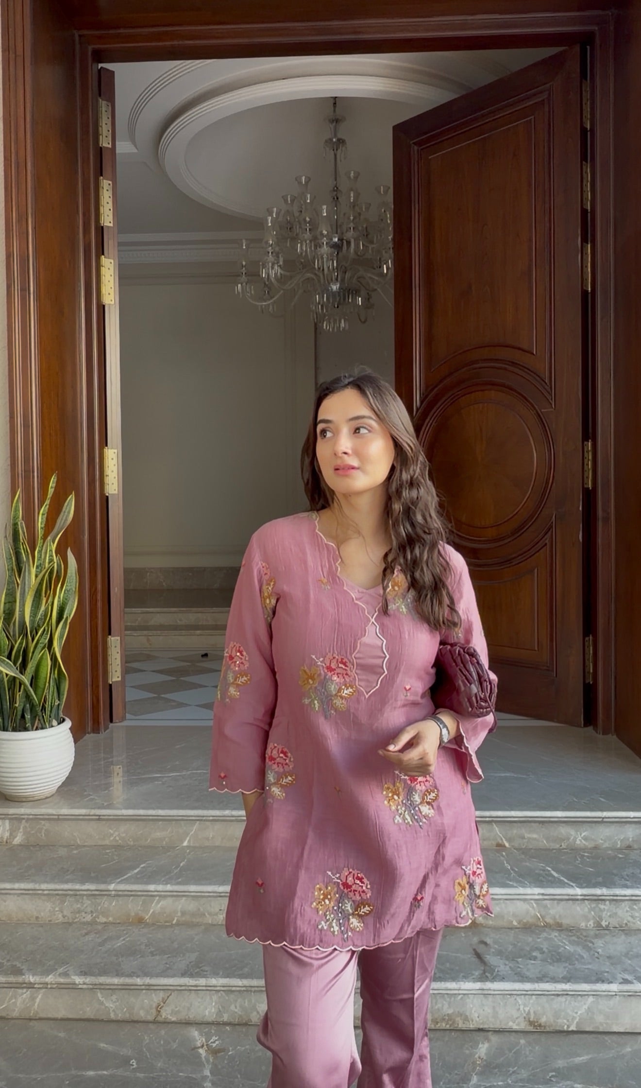 Woman in a pink embroidered outfit standing in a doorway with a chandelier in the background.