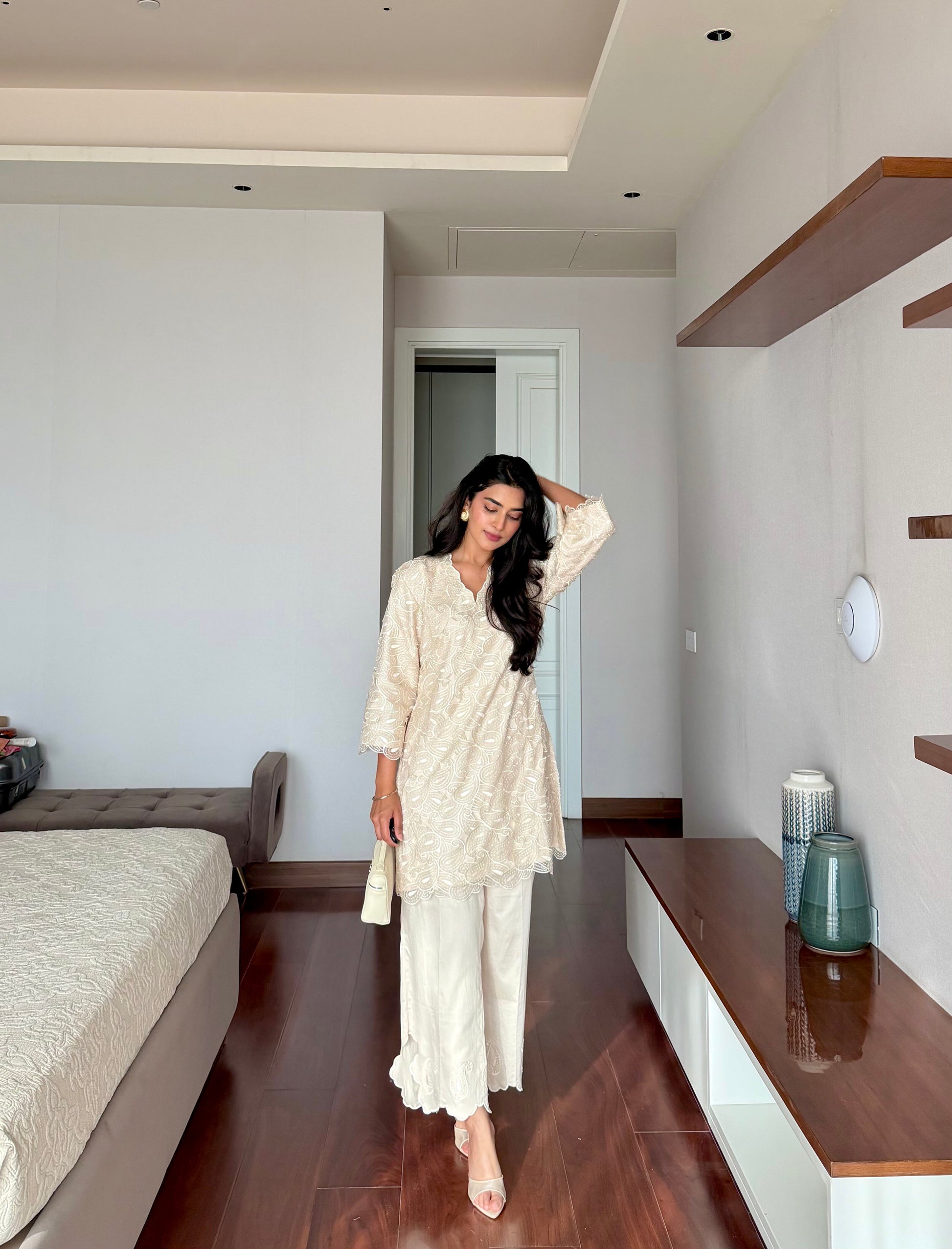 A woman stands in a modern bedroom wearing a cream-colored two-piece tunic and trouser set with a subtle floral pattern. The room features dark wood flooring, a bed on the left, and a built-in shelf on the right.