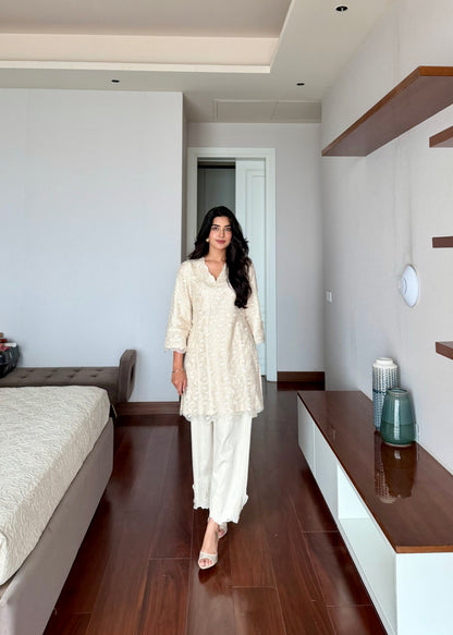 A woman modeling a cream-colored embroidered kurta and matching wide-leg trousers in a modern bedroom setting with dark wood floors.