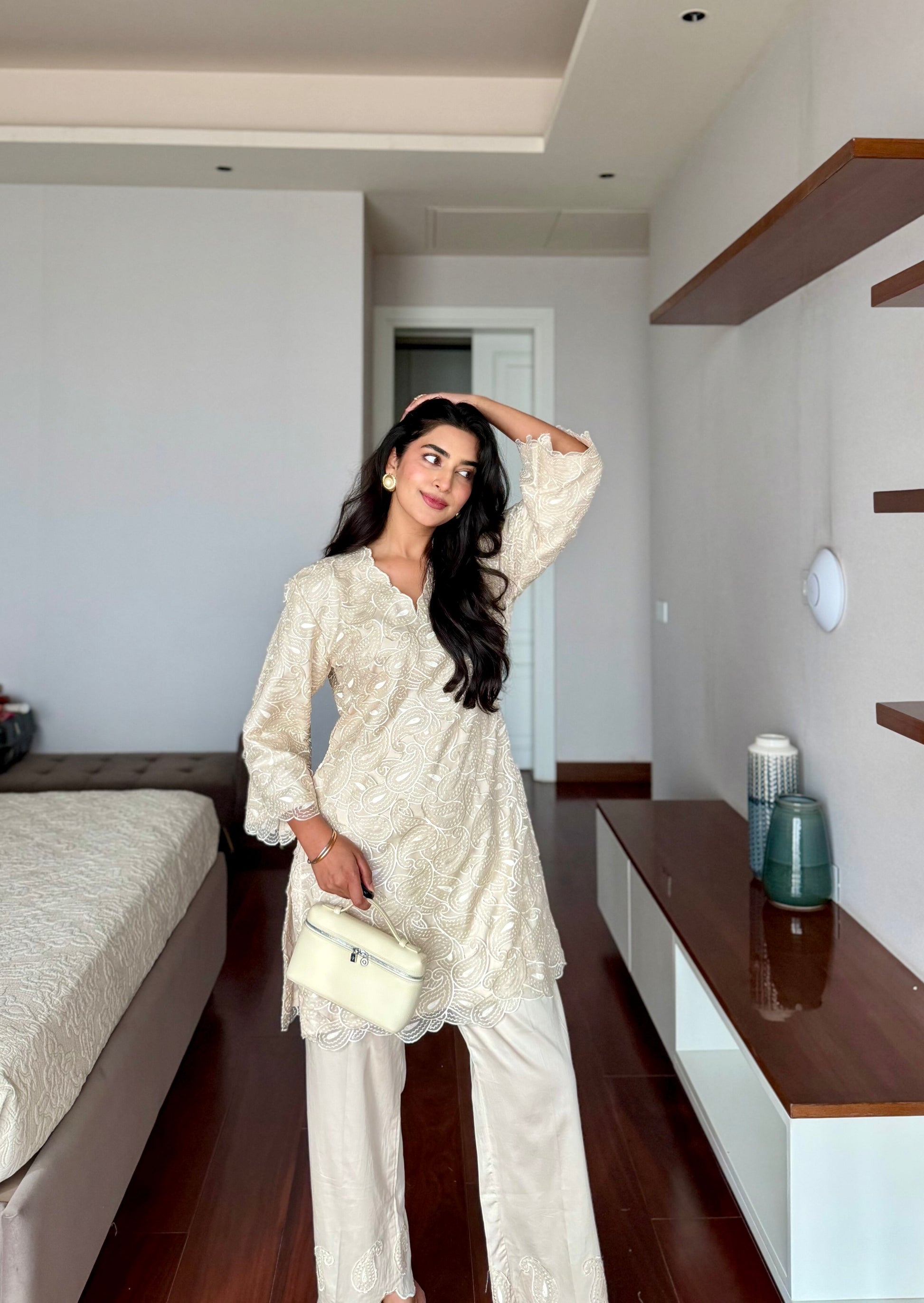 A woman posing in a cream-colored embroidered tunic and wide-leg trousers set, holding a matching cream handbag in a modern bedroom setting.