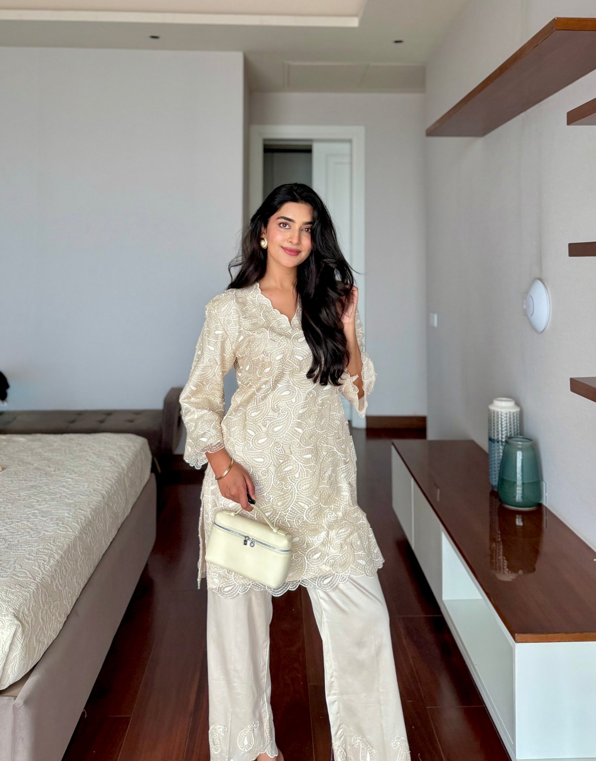 A woman stands in a bedroom wearing a cream-colored embroidered tunic and wide-leg trousers, holding a matching cream handbag. The room features dark wood floors, a bed on the left, and a white cabinet with wooden shelves on the right.