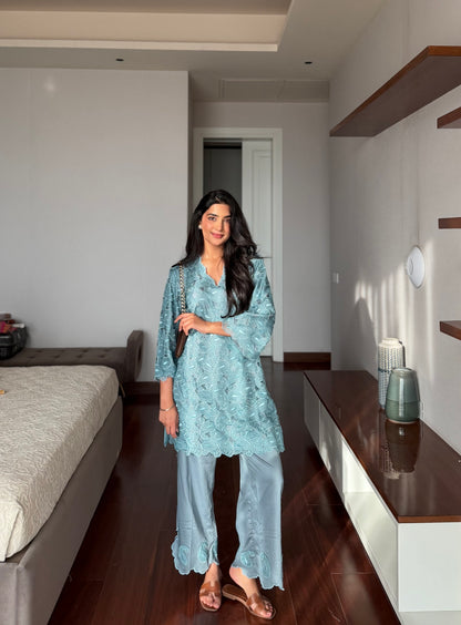 A woman stands in a modern bedroom wearing a light blue embroidered tunic with matching wide-leg pants and brown flat sandals.