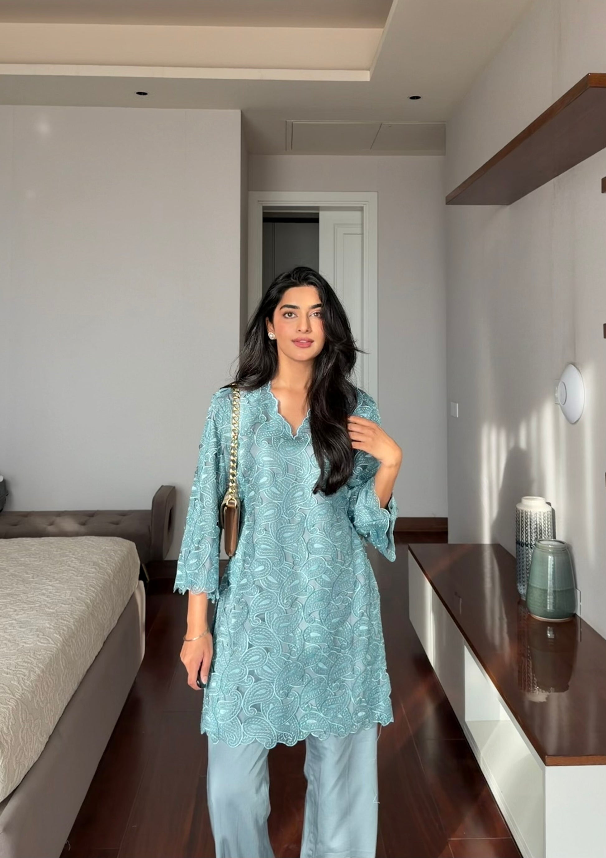 A woman wearing a light blue floral embroidered tunic top and matching light blue pants, standing in a modern bedroom setting.